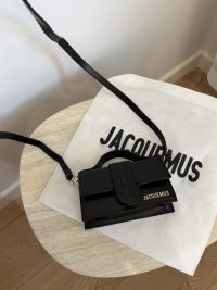 Black Jacquemus Le Bambino mini bag on table with branded dust bag.