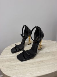 Black YSL Opyum sandals with gold logo heels on table.
